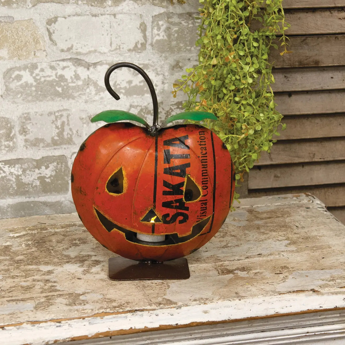 A decorative orange jack-o'-lantern metal lantern with green leaves and a black hanging hook, displayed on a weathered wooden surface against a rustic stone and wood wall backdrop.