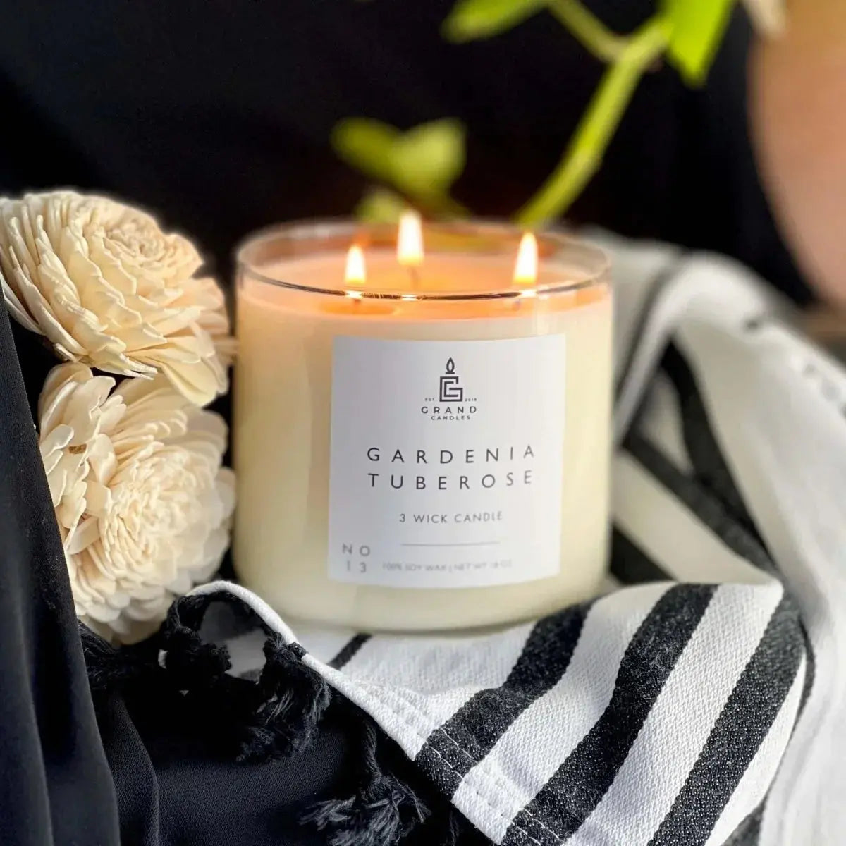 Gardenia Tuberose 3-wick soy candle with soft glow, styled with cream flowers and striped fabric for cozy home decor