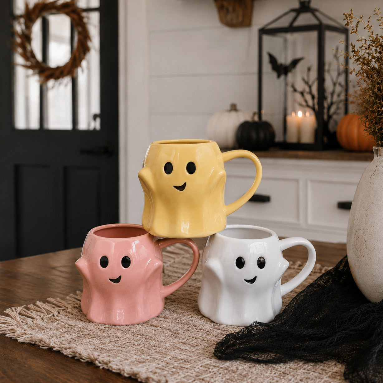 Three ghost-shaped mugs in pink, yellow, and white on a table with Halloween decorations in the background.