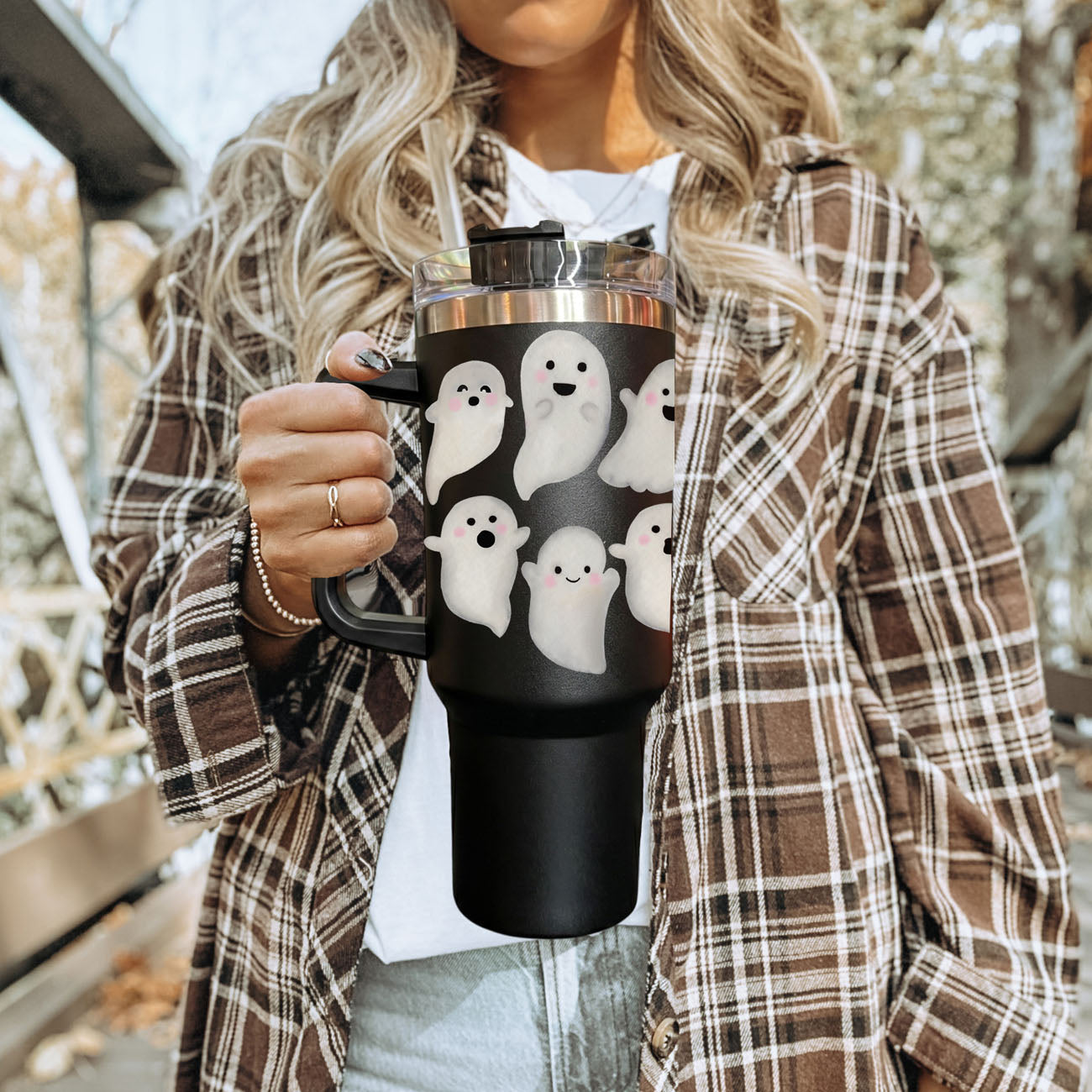 Person holding a black mug with ghost designs, wearing a plaid jacket outdoors.