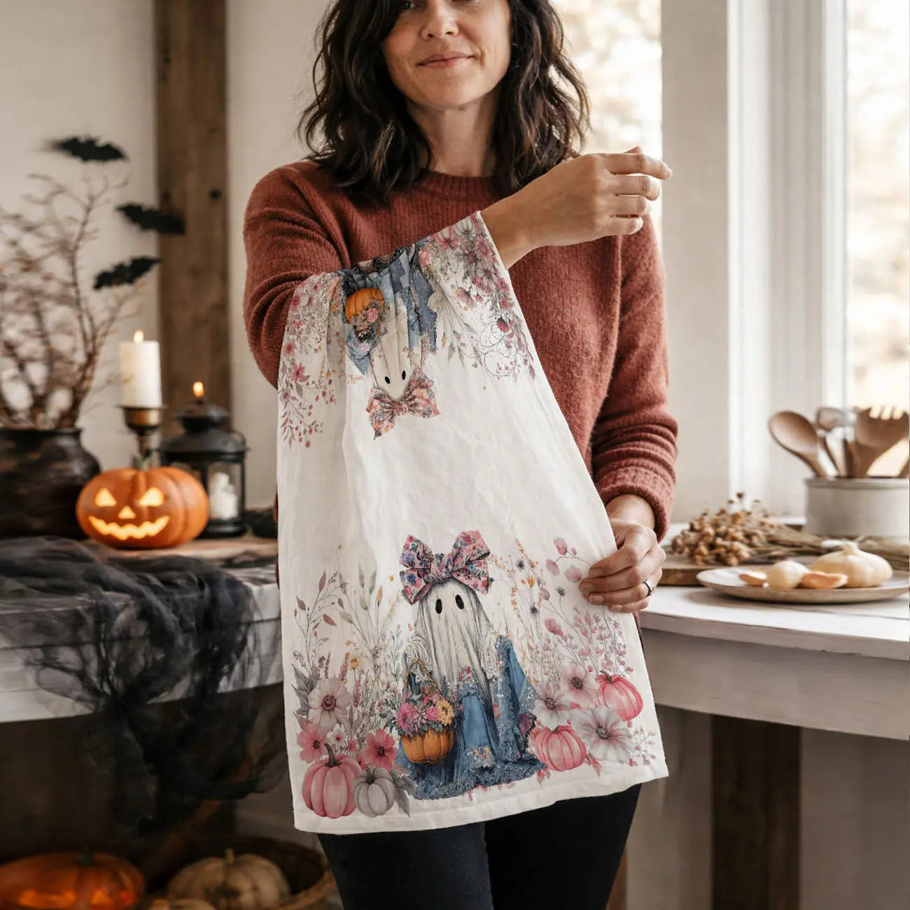 Woman holding a decorative kitchen towel with ghost and pumpkin design in a cozy kitchen.