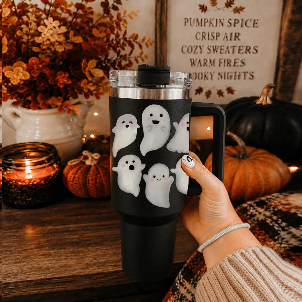 Black tumbler with ghost designs held by a hand in a cozy setting with pumpkins and candles.