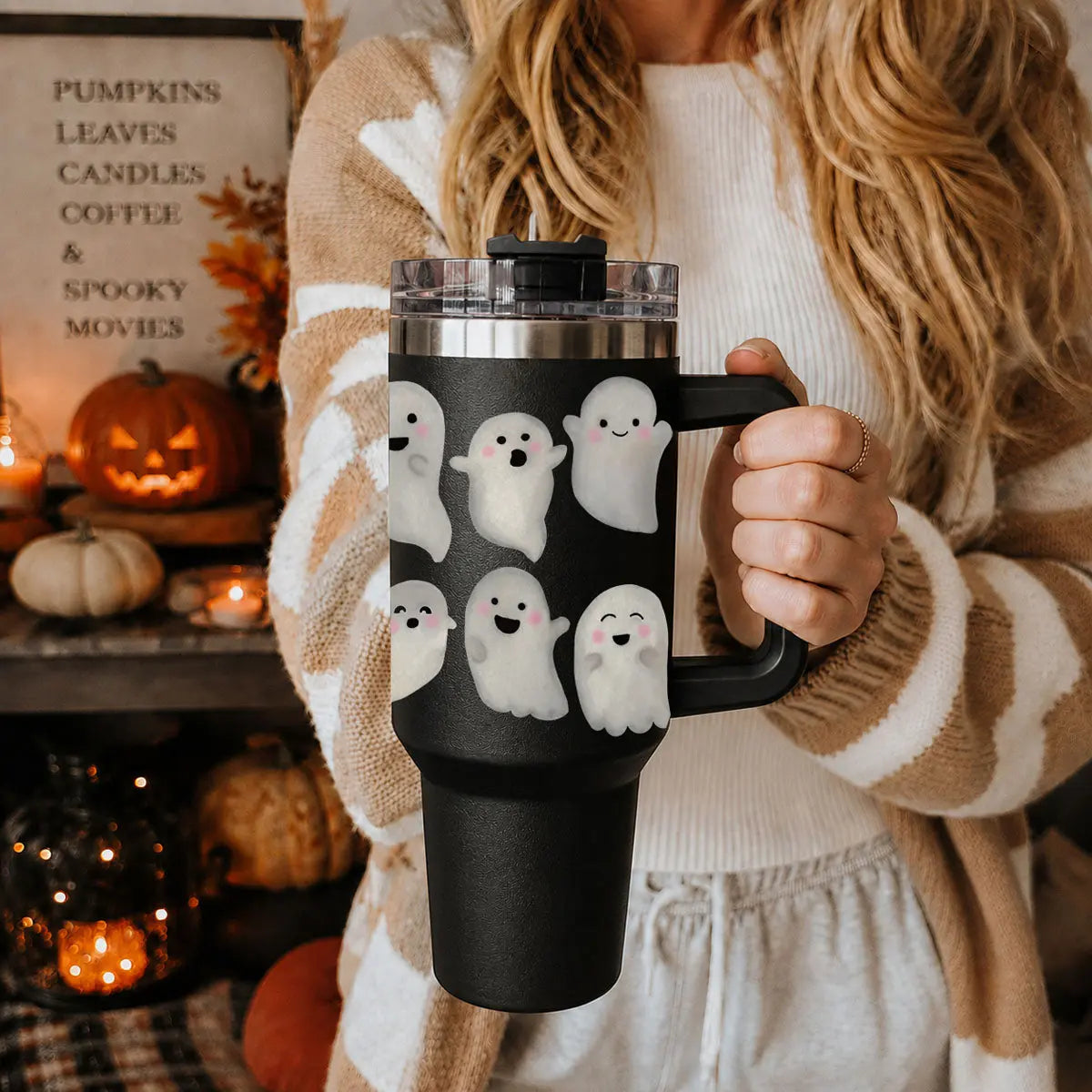 Person holding a black tumbler with ghost designs in a cozy indoor setting with Halloween decorations.