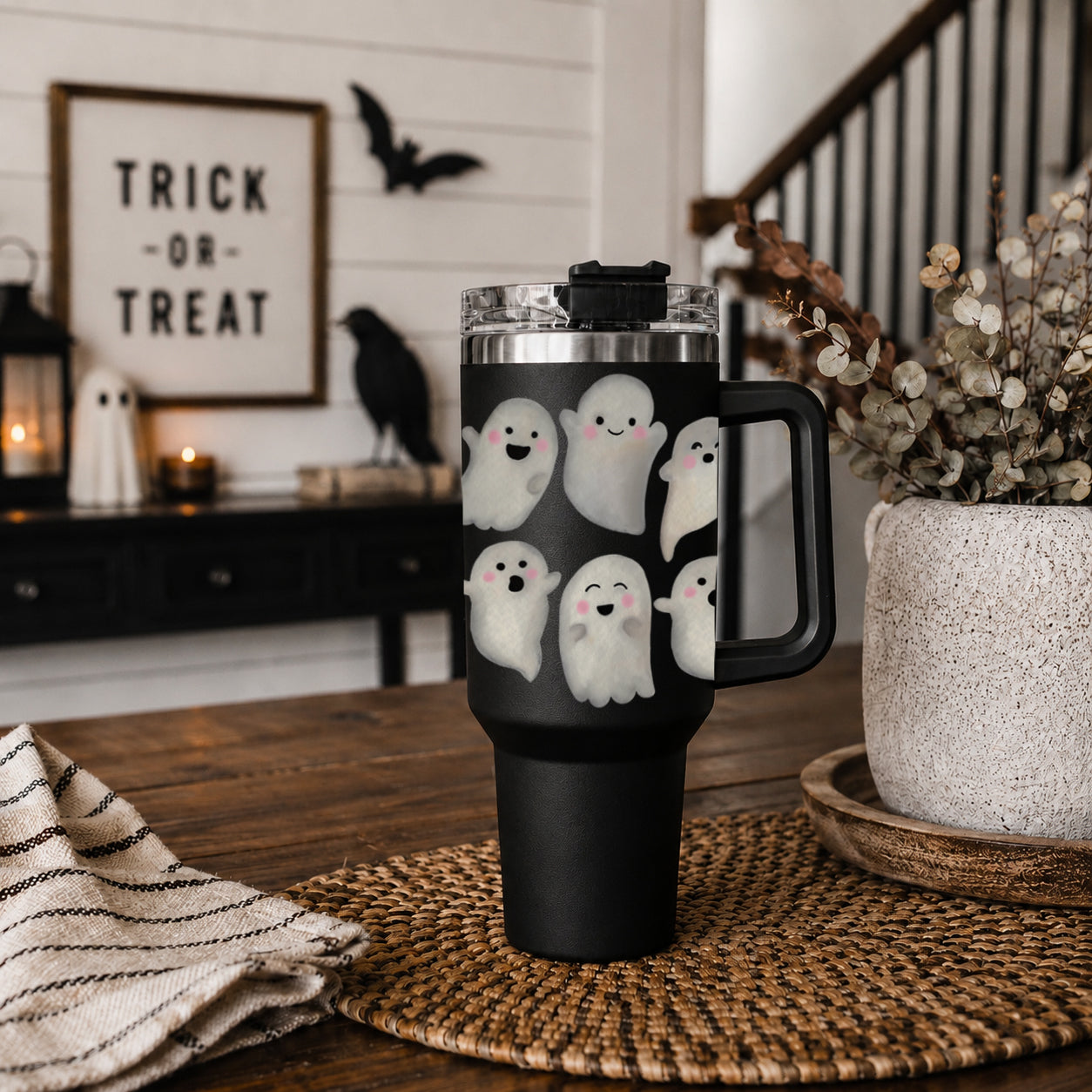 Black tumbler with ghost designs on a wooden table with Halloween decorations in the background.
