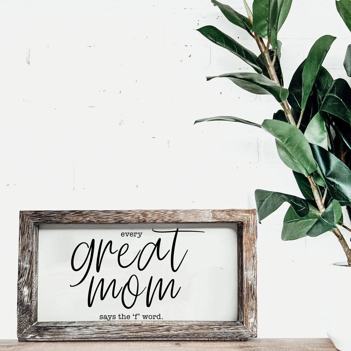 Framed print with 'Every great mom says the f-word' text on a white wall with a plant.
