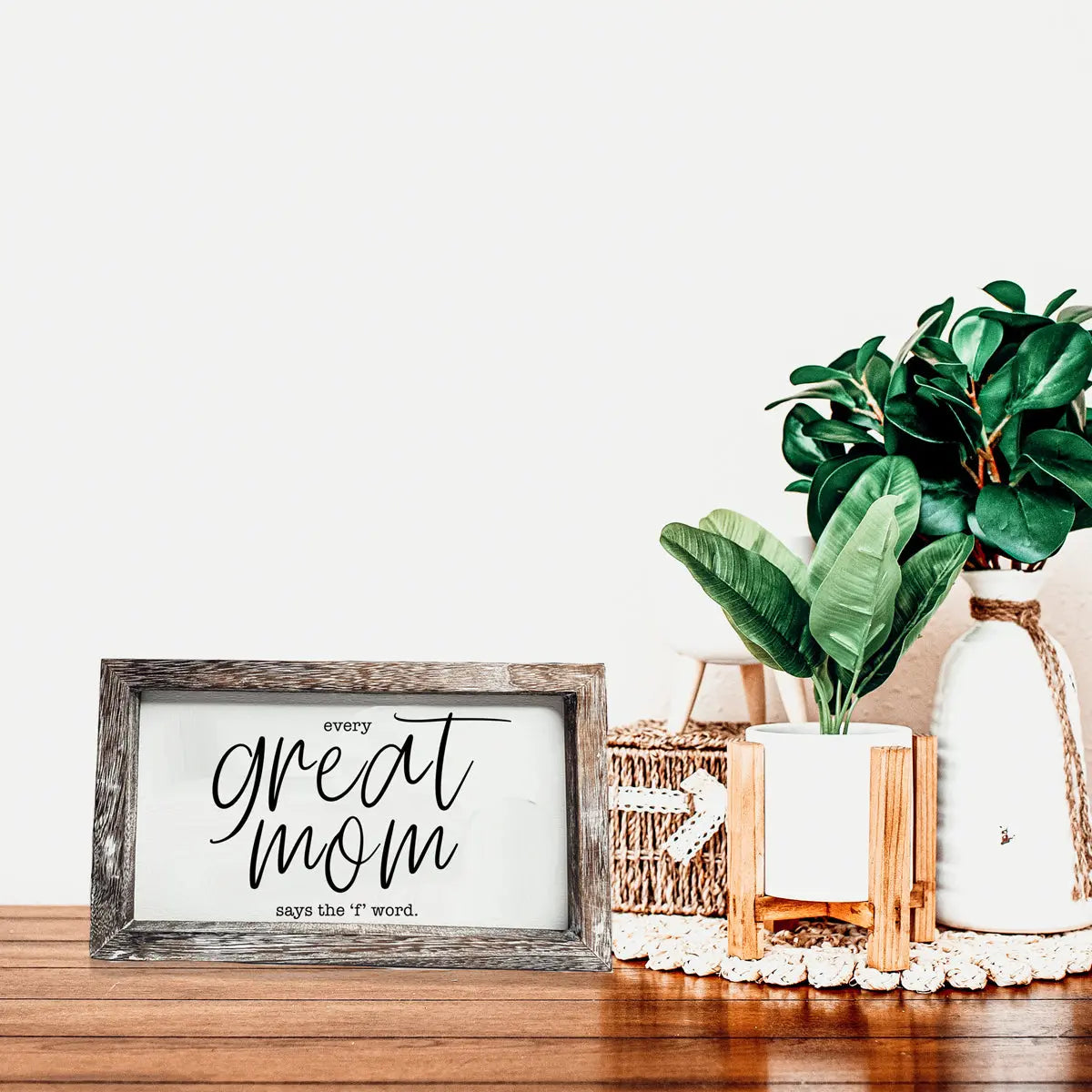 Decorative sign with 'every great mom says the t word' on a wooden surface with plants.