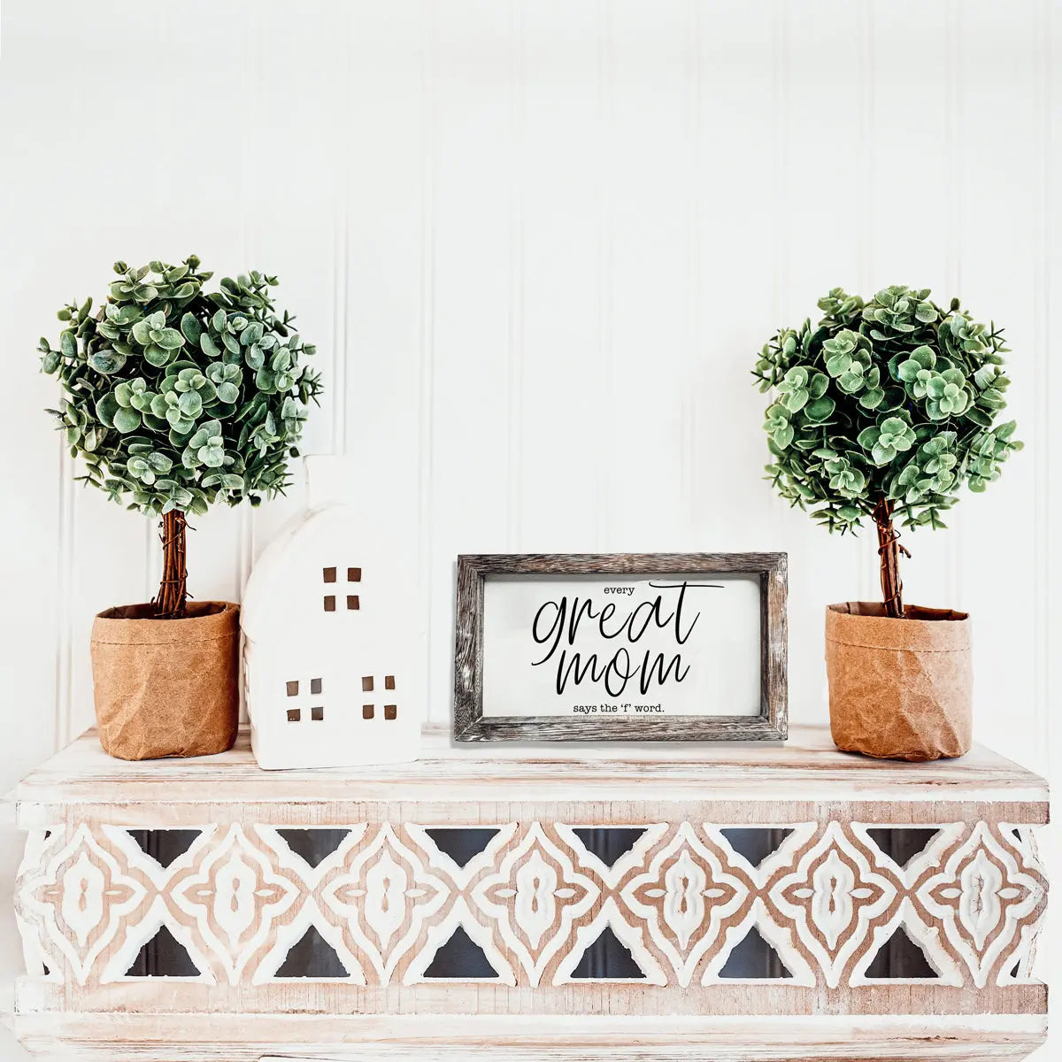 Decorative shelf with two potted plants, a white house-shaped object, and a framed sign on a white wall.