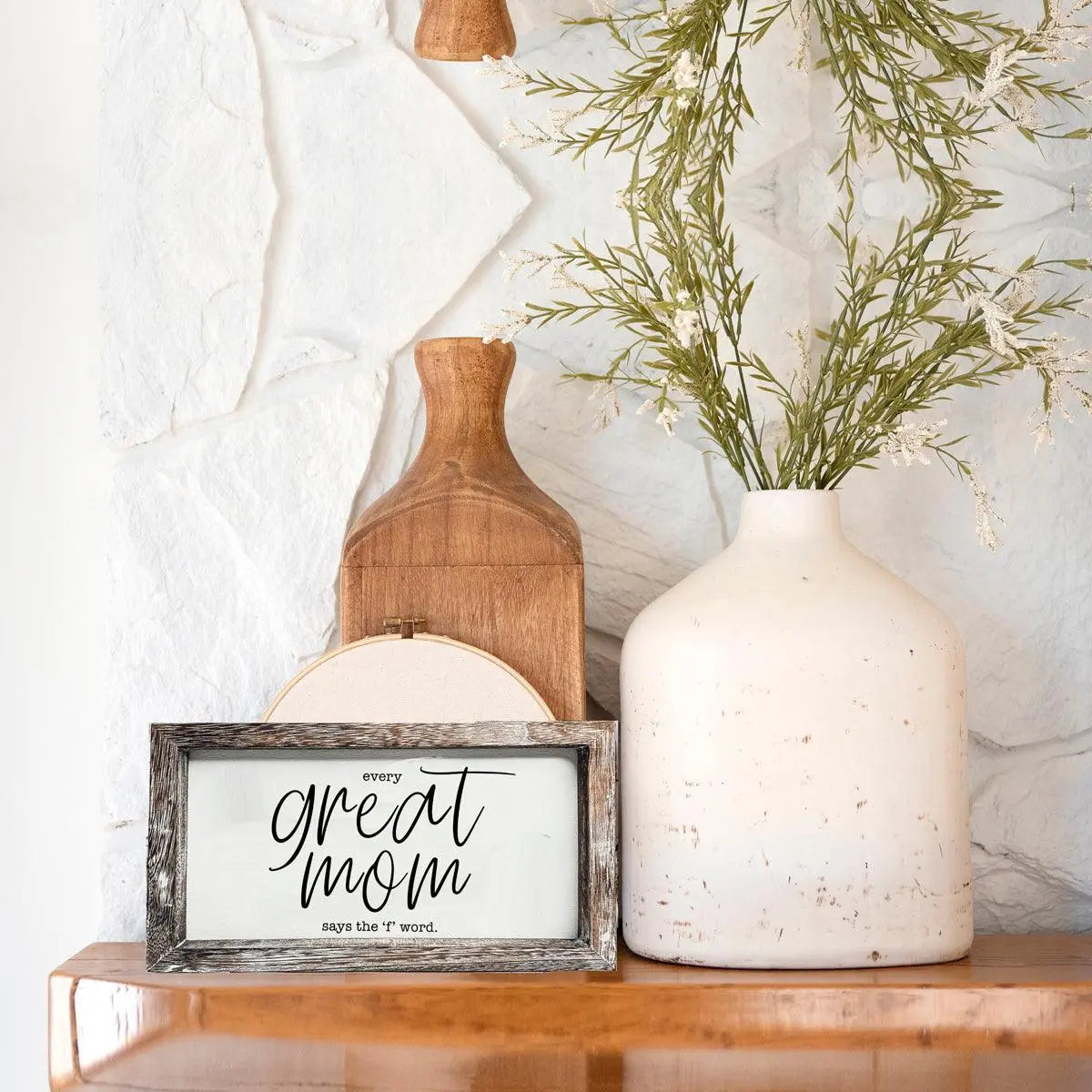 Decorative setup with a framed sign, wooden cutting board, and white vase with greenery on a marble surface.