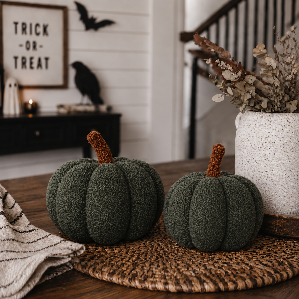 Two green textured pumpkins on a woven mat with Halloween decorations in the background.