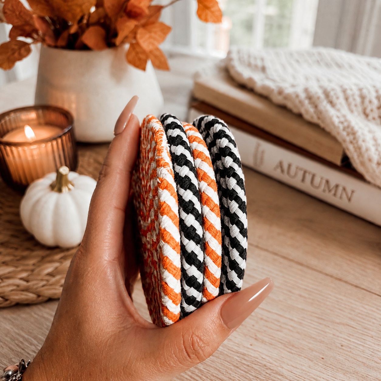 Hand holding three striped coasters with a cozy autumn setting in the background.