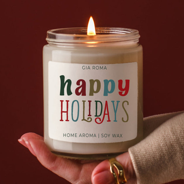 Candle with 'happy holidays' label held by a hand against a dark background