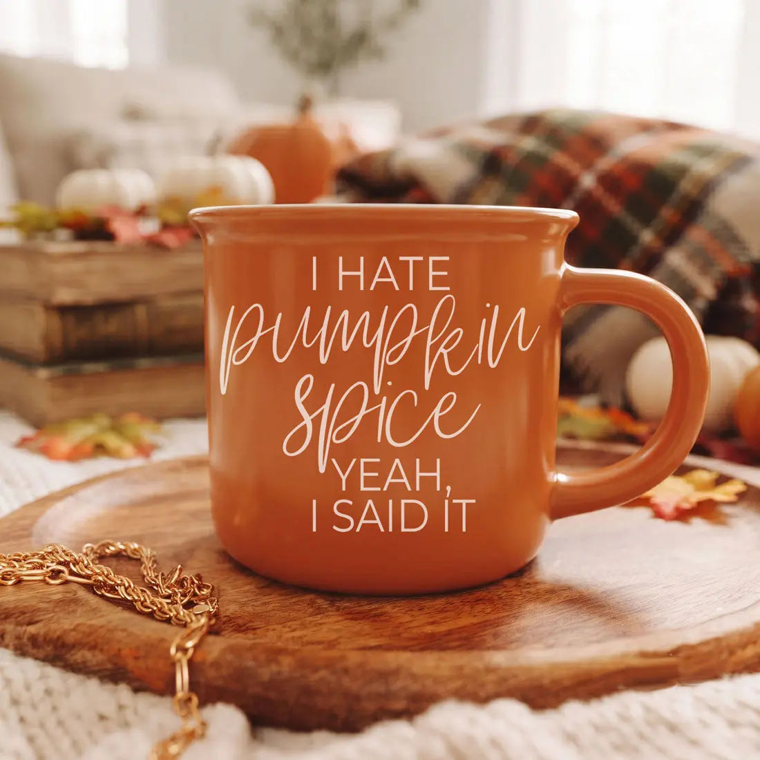 An orange ceramic mug with white text reading 'I HATE pumpkin spice YEAH, I SAID IT' sits on a wooden board in a cozy autumn setting with pumpkins, a plaid blanket, and a gold chain.