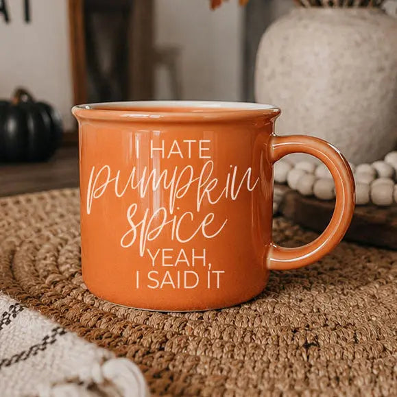 An orange ceramic mug with white text reading 'HATE pumpkin spice YEAH, I SAID IT' sits on a woven placemat in a cozy indoor setting with autumn decor blurred in the background.