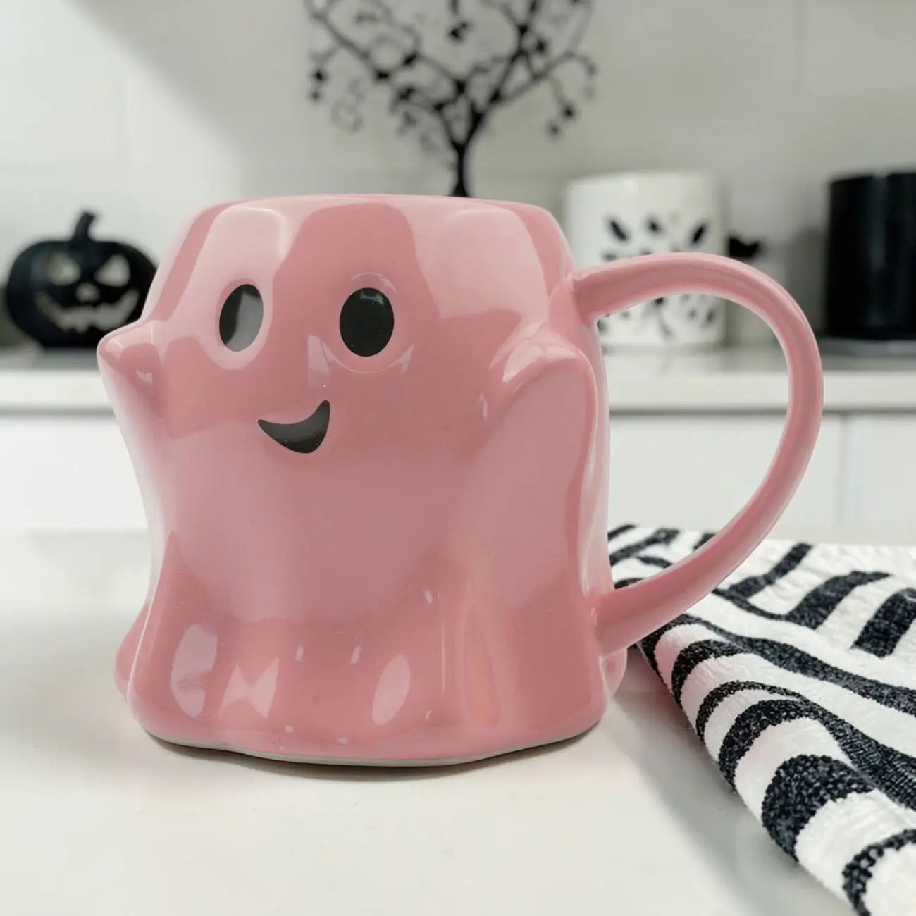 Pink ghost-shaped mug on a surface with Halloween decorations in the background