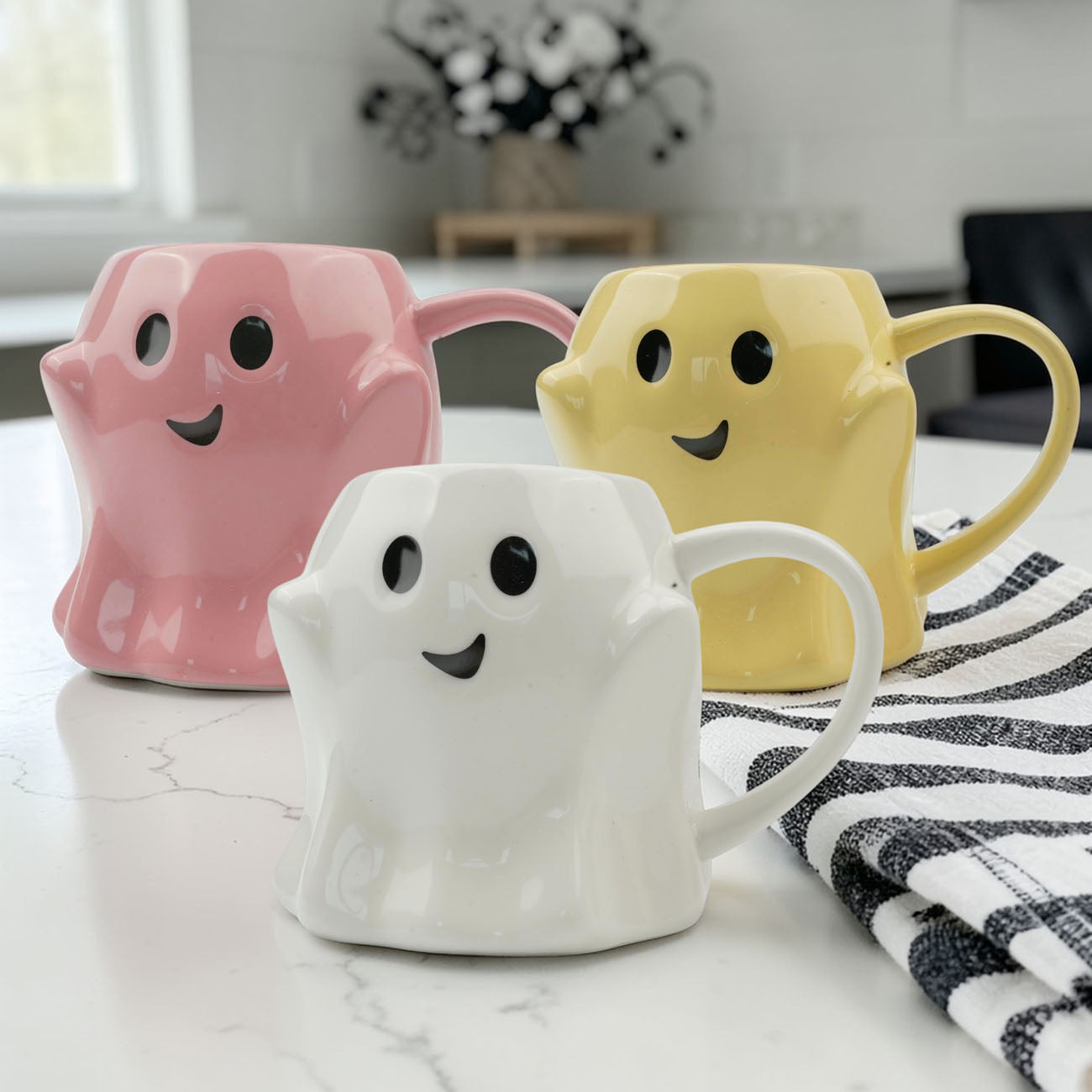 Three ghost-shaped mugs in pink, white, and yellow on a marble surface with a striped towel.