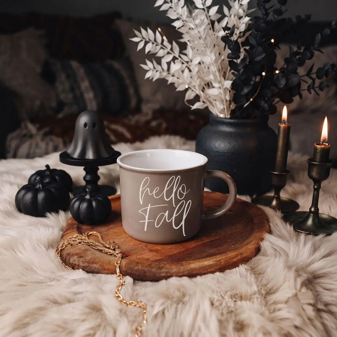 A gray ceramic mug with white text reading 'autumn leaves pumpkins please' sits on a round wooden tray. The mug is styled in a cozy fall setting with black pumpkins, a black ghost-shaped candle holder, lit candles, and dried white and black foliage on a fluffy cream rug.