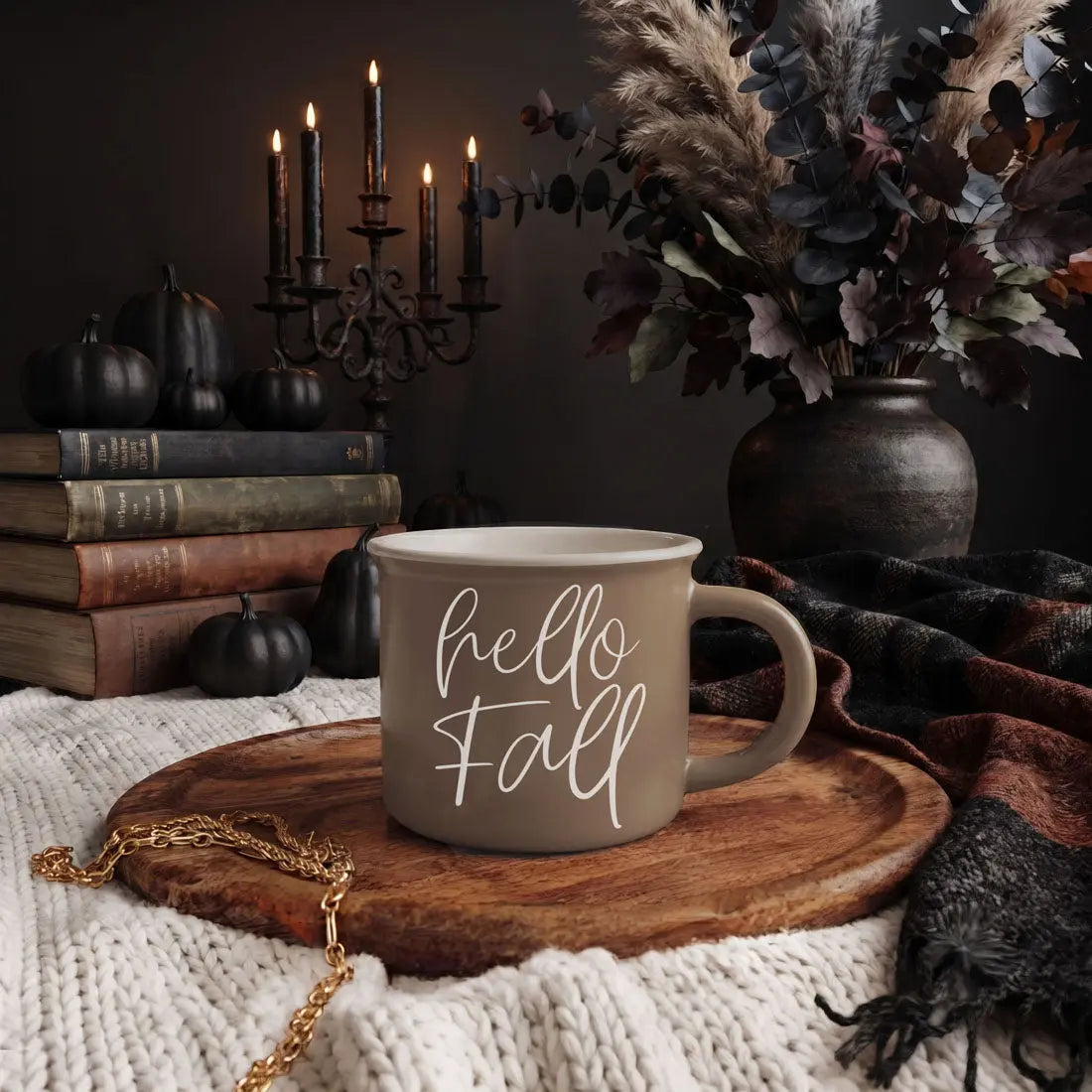 A beige ceramic mug with white script text reading 'autumn LEAVES pumpkins PLEASE' sits on a wooden tray in a styled fall scene with candles, books, pumpkins, dried flowers, and a knit blanket.