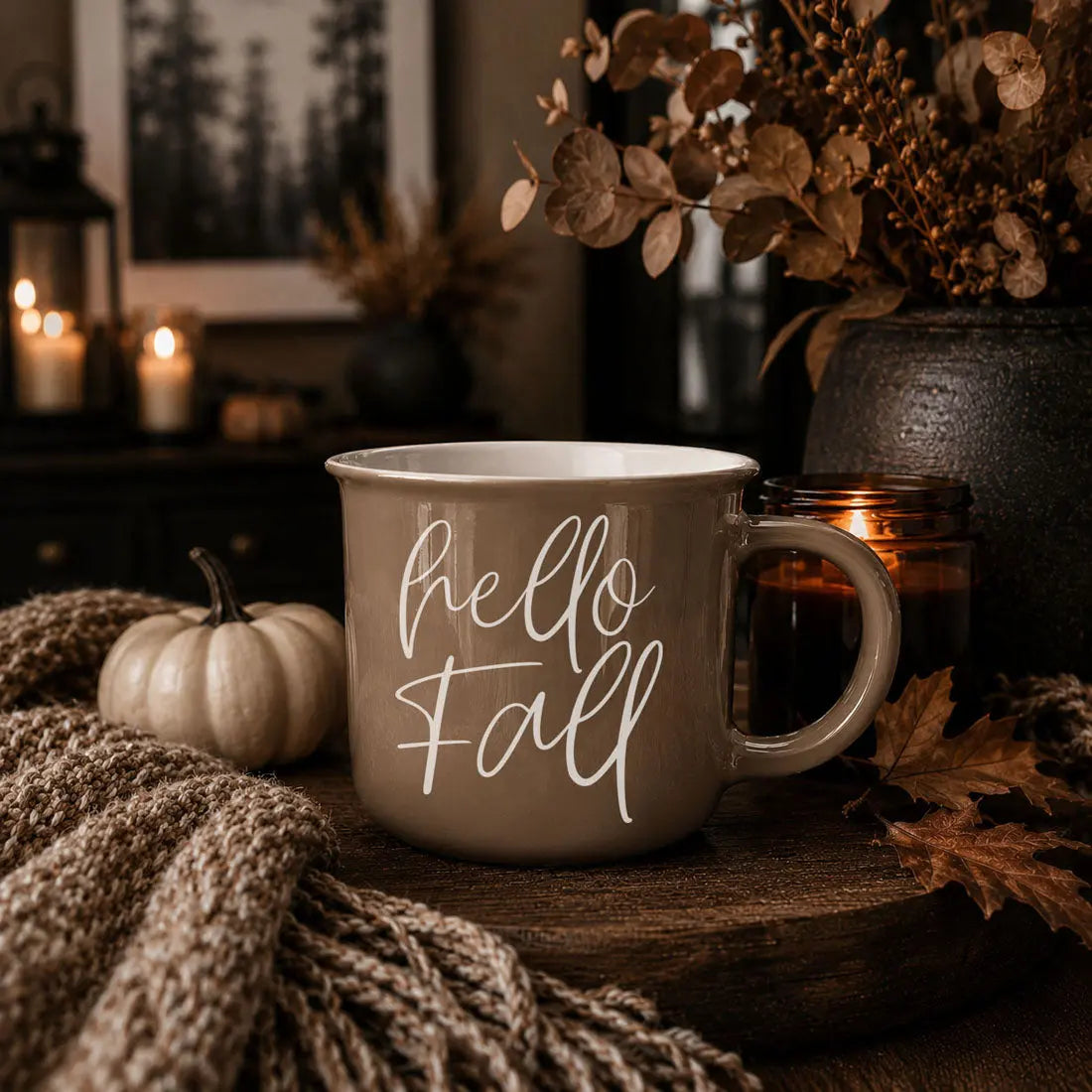 A ceramic mug with the phrase 'autumn LEAVES pumpkins PLEASE' in white script and block lettering, styled on a wooden surface with a knit blanket, a small pumpkin, a candle, and dried foliage in a warm autumn setting.
