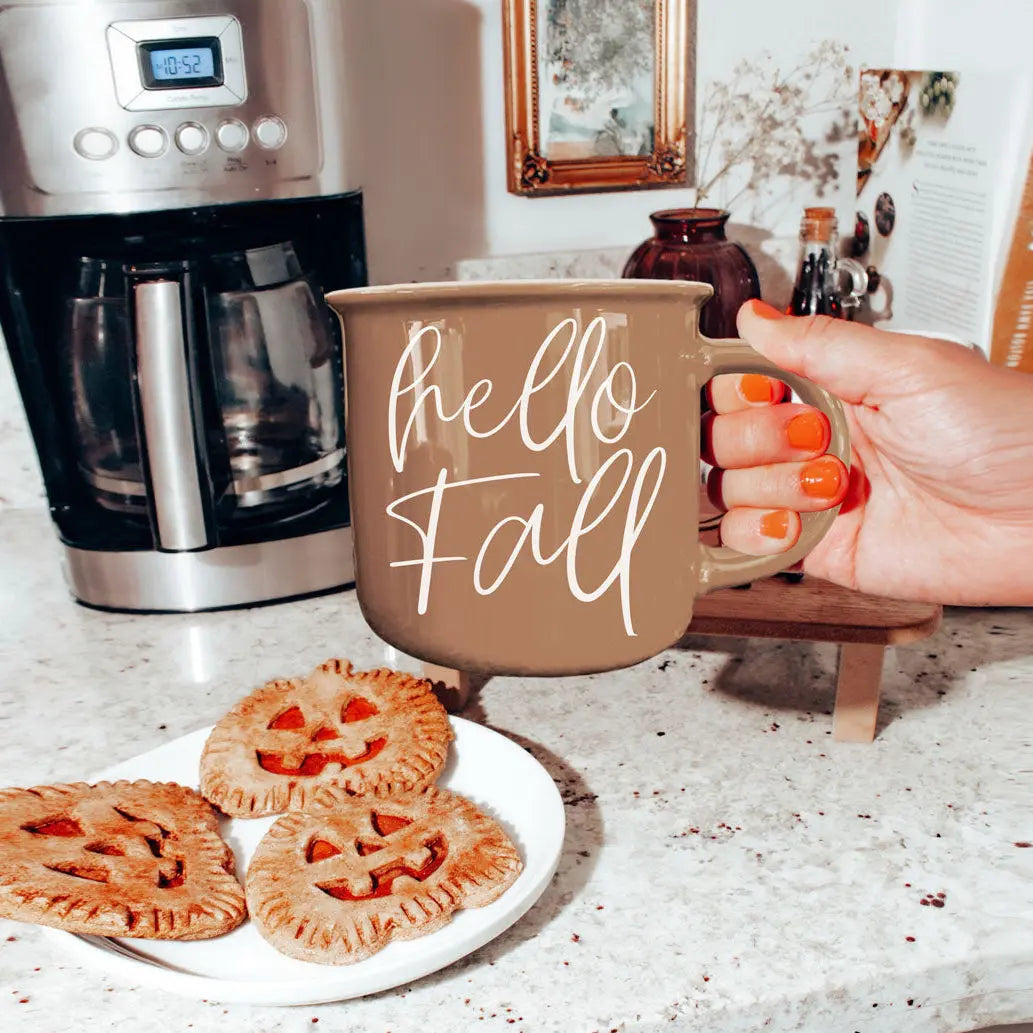 Hand holding a 'hello Fall' mug with pumpkin pies on a plate and a coffee maker in the background.