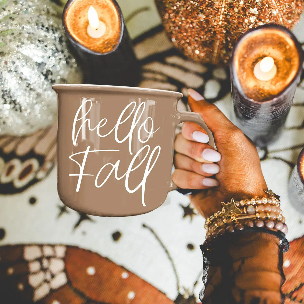 Hand holding a 'hello fall' mug with candles and decorative items in the background