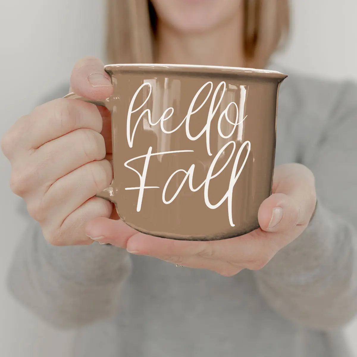 A person holding a brown enamel-style mug with the words 'hello Fall' written in white script, shown against a soft neutral background.