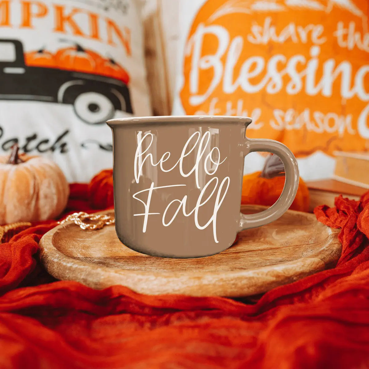 Brown mug with 'hello Fall' text on a wooden coaster with pumpkins and autumn-themed decor in the background.
