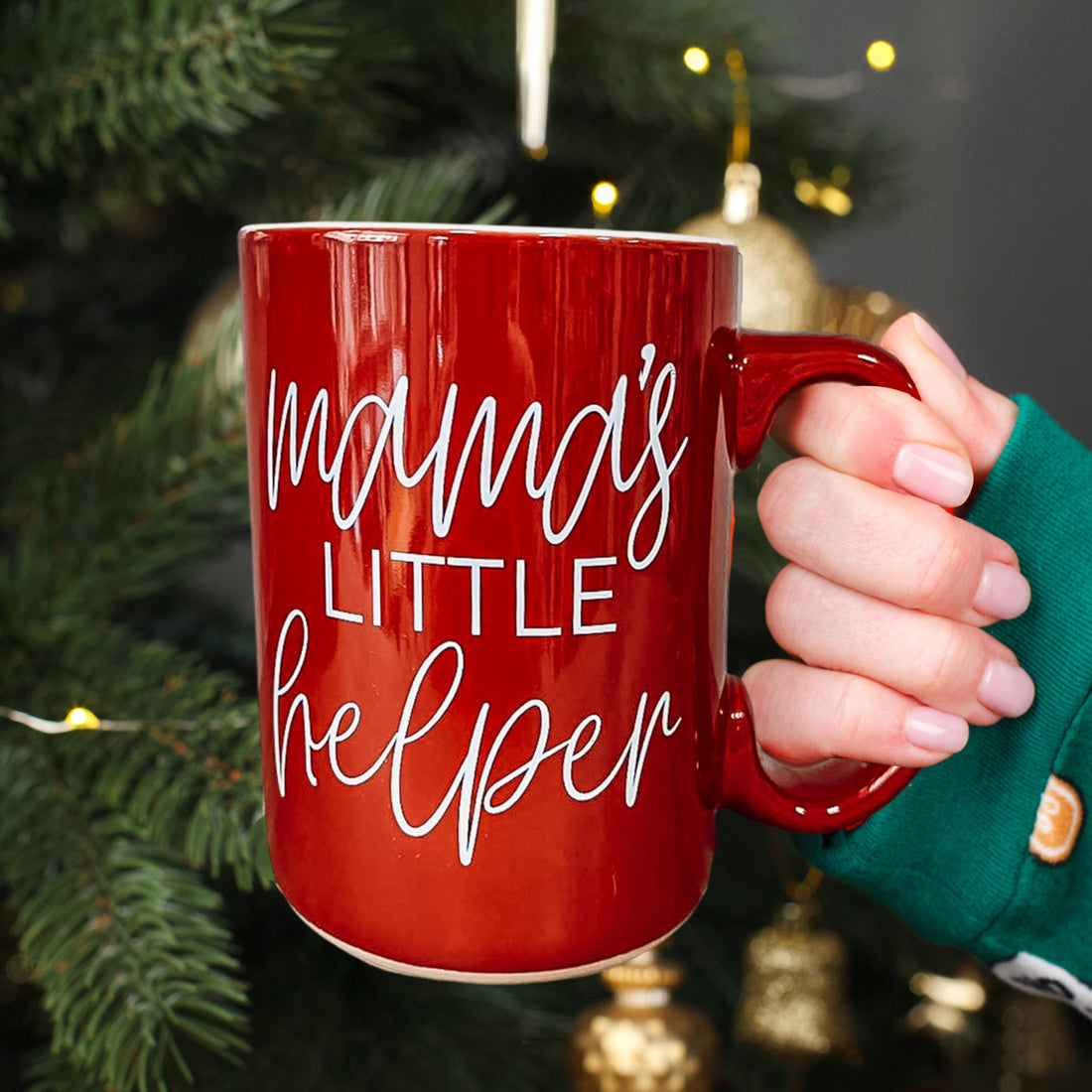 Red mug with 'mama's little helper' text held by a hand in front of a decorated Christmas tree.