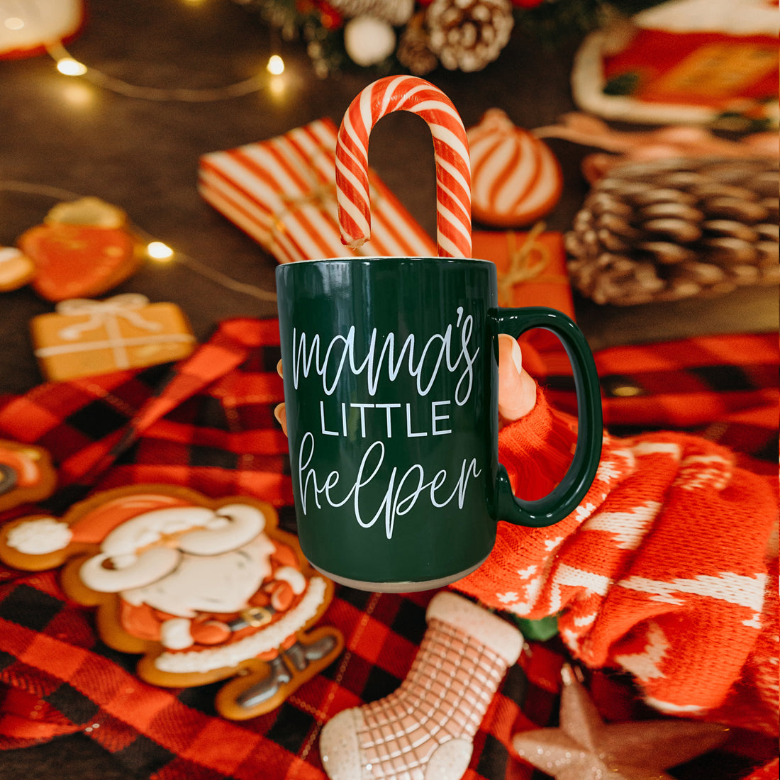 Green mug with 'mama's little helper' text, candy cane handle, surrounded by festive decor.