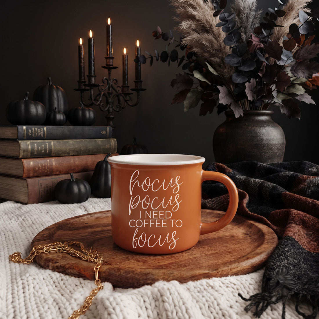 An orange ceramic coffee mug with white text reading 'hocus focus I NEED COFFEE TO focus' sits on a round wooden tray. The scene is styled in a cozy autumn setting with a black candelabra, books, pumpkins, a dark vase of dried foliage, and a plaid throw blanket.