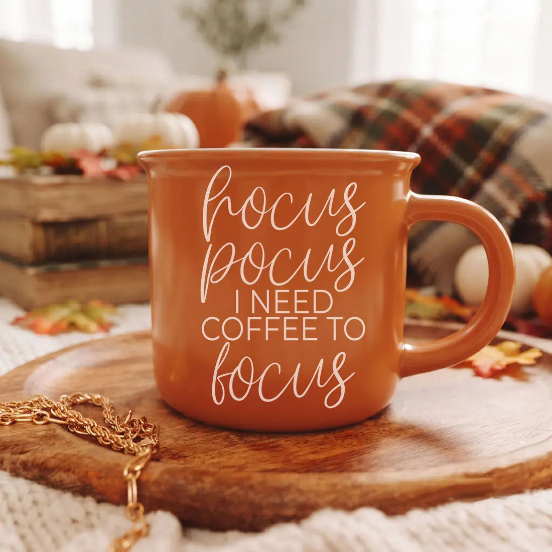 An orange ceramic mug with white text reading 'focus pocus I NEED COFFEE TO focus' sits on a wooden tray in a cozy autumn-themed setting with pumpkins, a plaid blanket, and a gold chain.