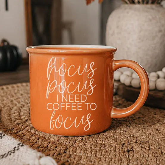 An orange ceramic coffee mug with white text reading 'focus pocus I NEED COFFEE TO focus' sits on a woven placemat in a cozy, softly blurred home setting.