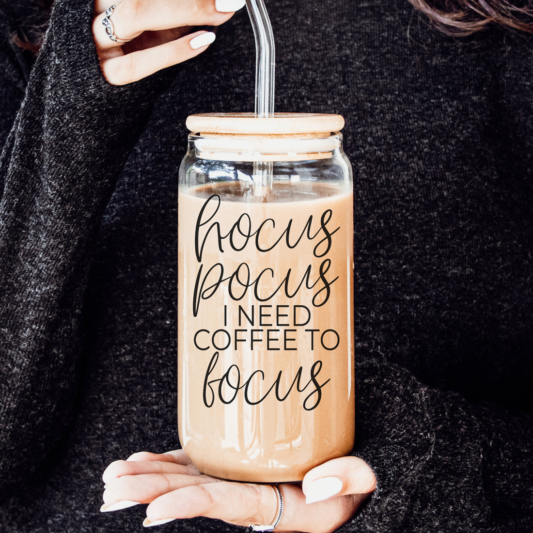Glass jar with straw lid filled with coffee, featuring text 'hocus pocus I need coffee to focus', held by a person.