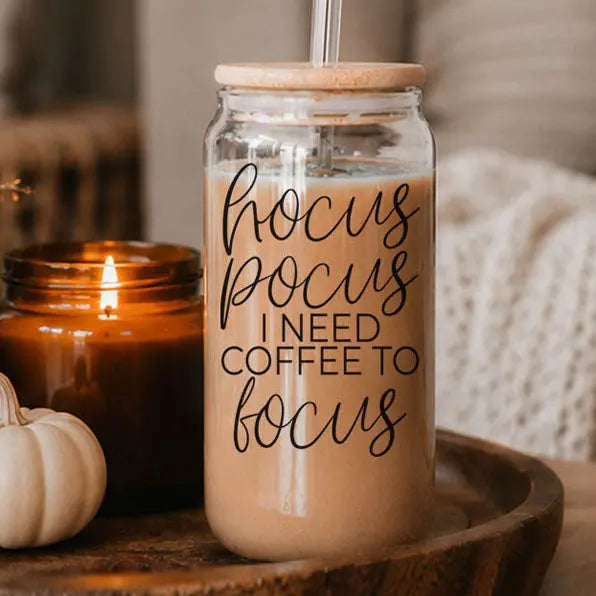 Glass jar with straw and wooden lid filled with a brown liquid, featuring text, on a wooden surface with a candle and pumpkin in the background.