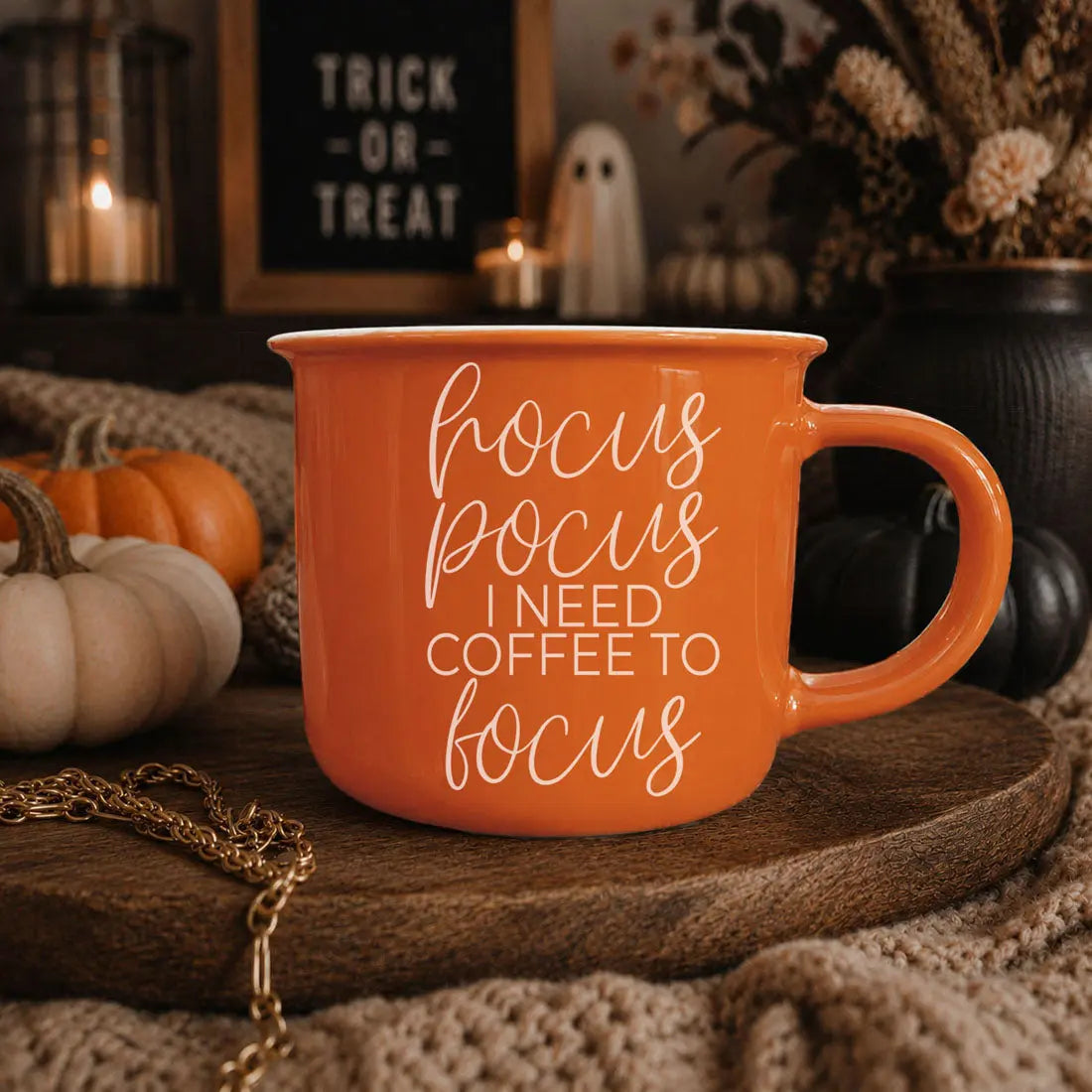 An orange ceramic mug with white text reading 'hocus pocus I NEED COFFEE TO focus' sits on a wooden tray in a cozy autumn-themed setting with pumpkins, candles, and a knitted blanket.