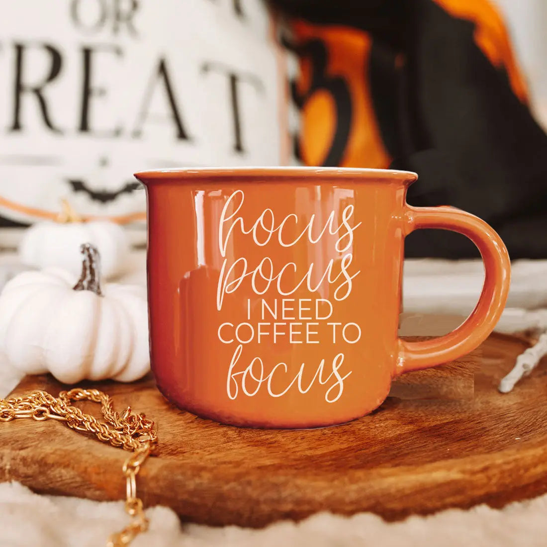 An orange ceramic mug with white text reading 'hocus pocus I NEED COFFEE TO focus' sits on a wooden tray with fall-themed props including small pumpkins, a gold chain, and a blurred sign in the background.