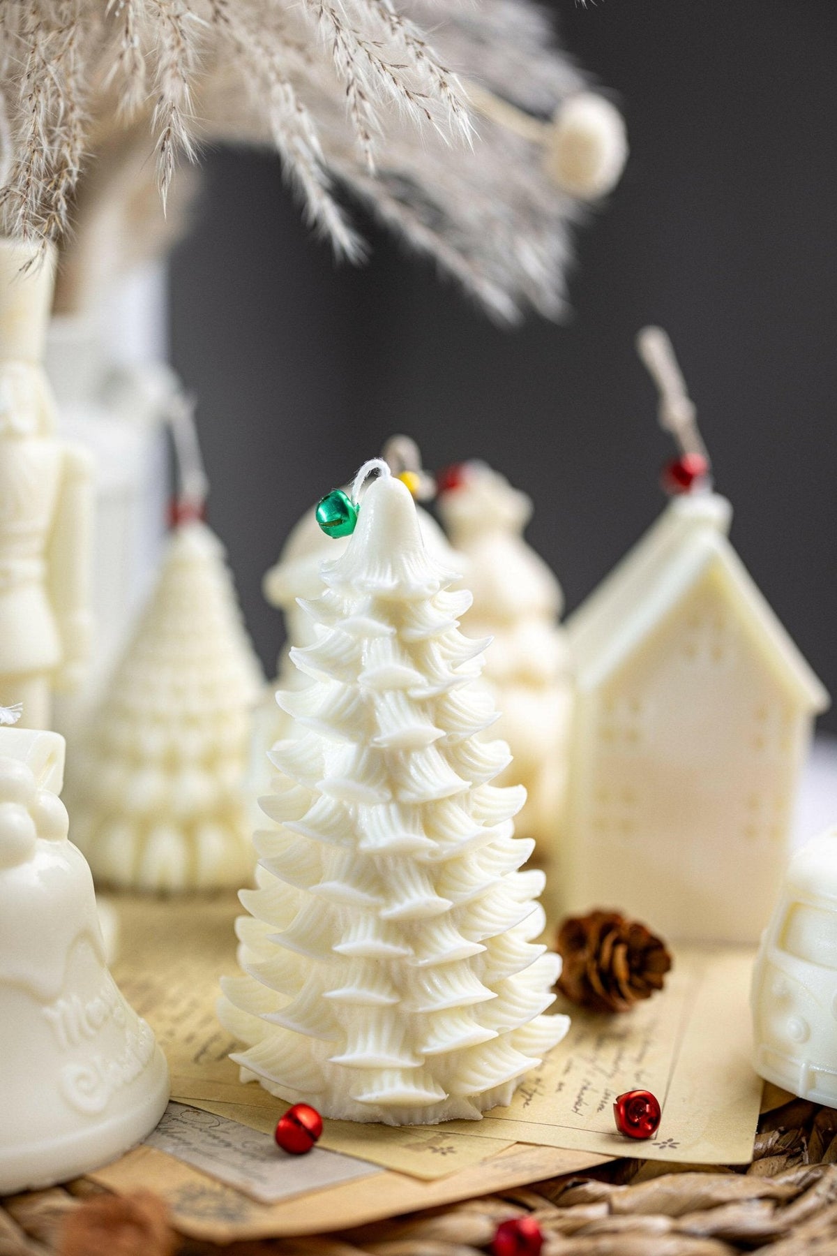 A white Christmas tree-shaped candle with a textured, layered design, decorated with small colorful ornaments, displayed on a rustic surface with other holiday-themed candles and decorations.