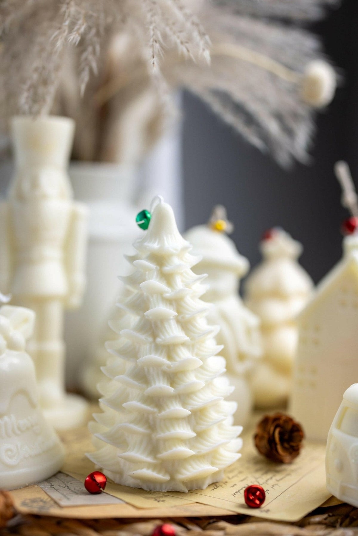 A white Christmas tree-shaped candle with a small green ornament on top, styled on a tabletop with other white holiday candles and dried grass in the background.