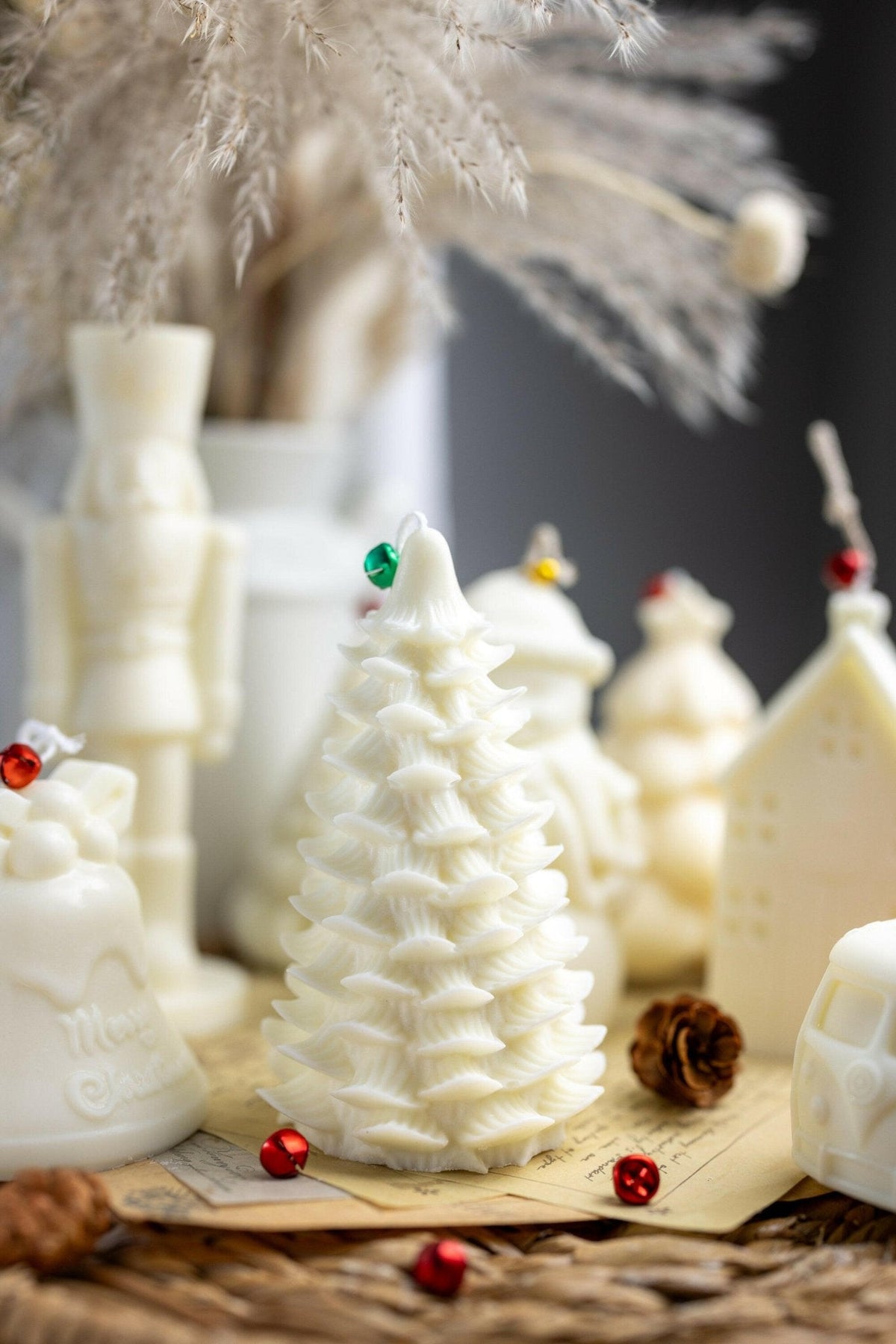 A collection of white Christmas-themed candles, including a textured tree-shaped candle, a nutcracker, a bell, and a house, styled on a rustic woven mat with dried grass and small red ornaments.