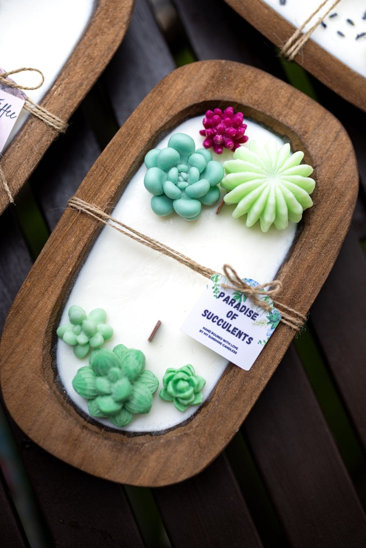 A wooden tray candle featuring a white wax base decorated with sculpted green succulents and a small pink flower, wrapped with twine and a tag reading 'Paradise of Succulents'.