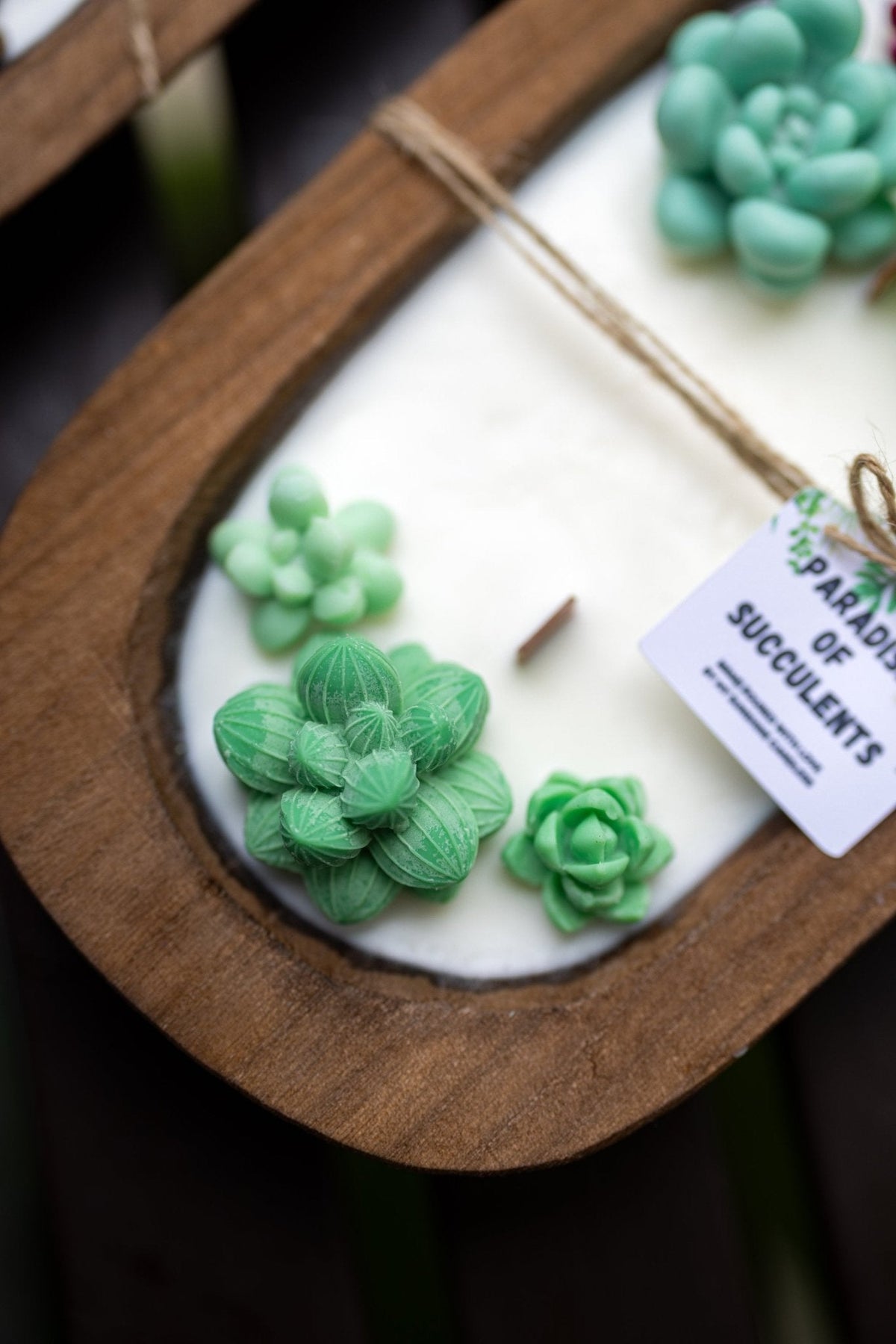 A close-up of a wooden tray containing a white candle with green succulent decorations and a small paper tag that reads 'PARDON ME SUCCULENTS'.