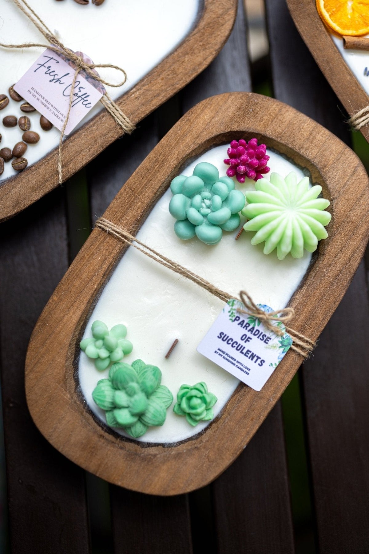 An oval wooden tray holding a white candle with sculpted succulent and flower decorations in mint green, light green, and magenta. A small paper tag is tied to the front with twine.