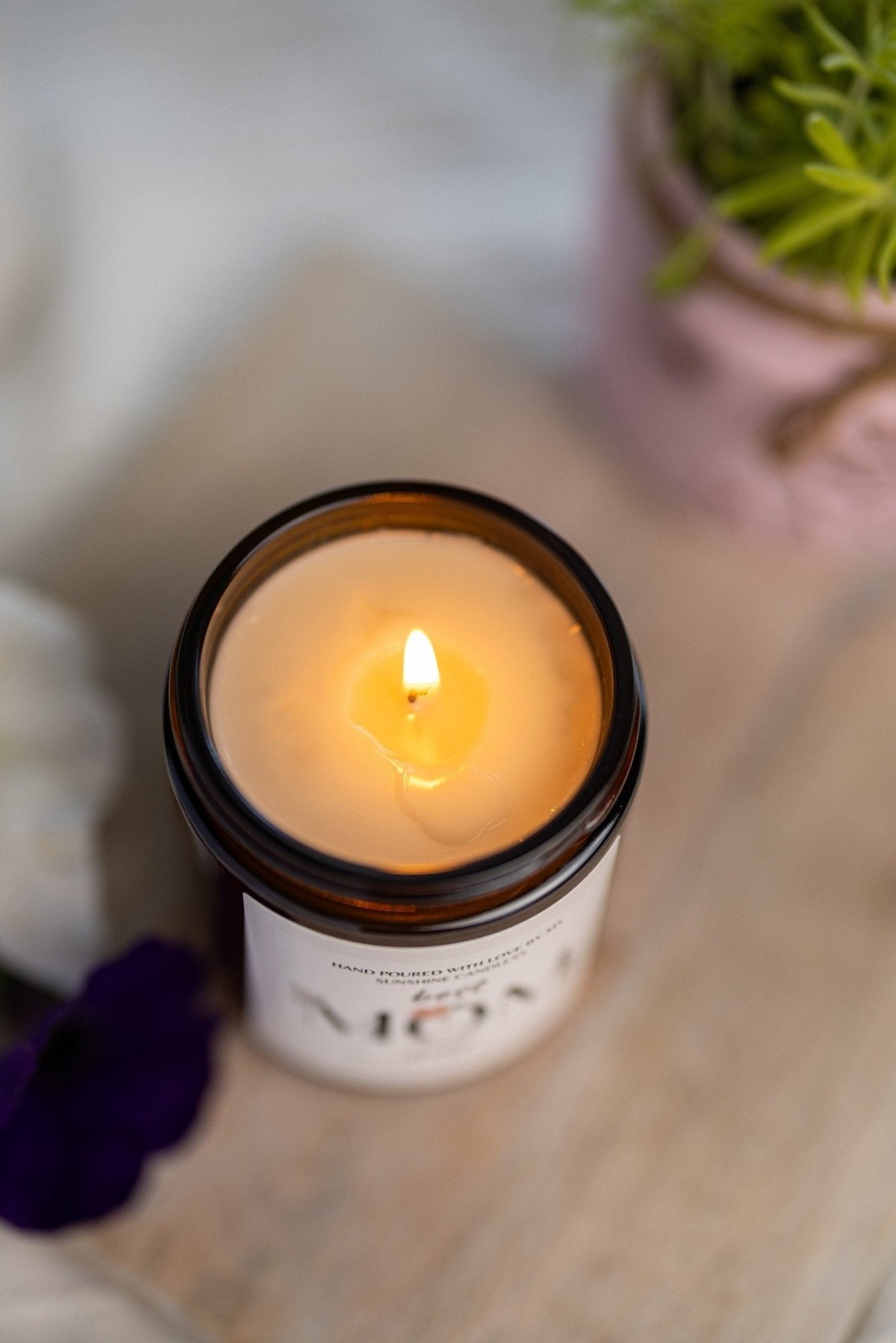 A lit candle in an amber glass jar with a white label, photographed on a light wooden surface with a blurred pink planter and green plant in the background.