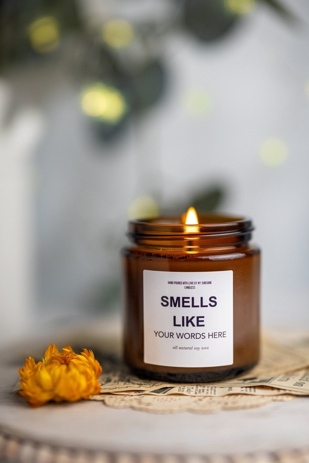 An amber glass jar candle with a lit flame sits on a rustic surface with a white label reading 'SMELLS LIKE YOUR WORDS HERE.' A small yellow flower rests beside it, and the background is softly blurred with warm lights and greenery.
