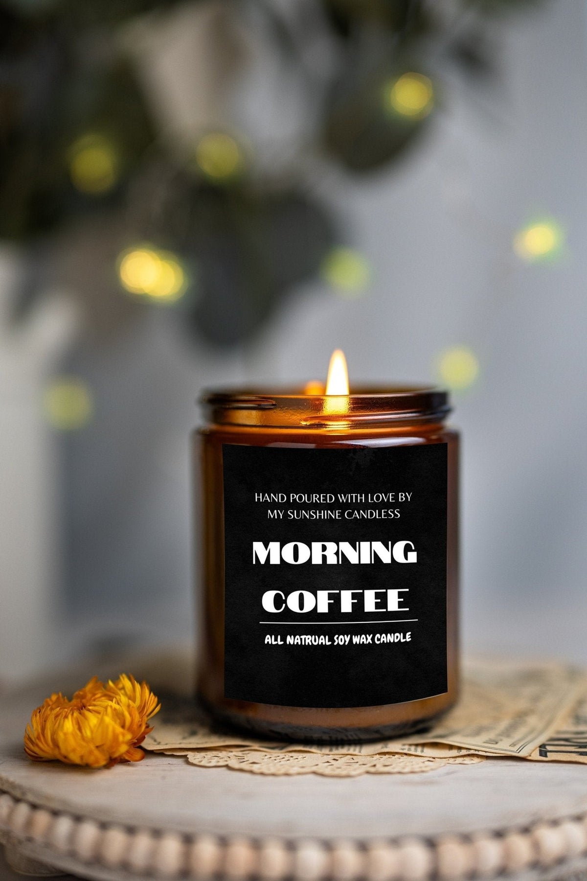 A lit amber glass candle jar labeled 'MORNING COFFEE' sits on a decorative surface with a small yellow flower beside it and a softly blurred background with warm lights.