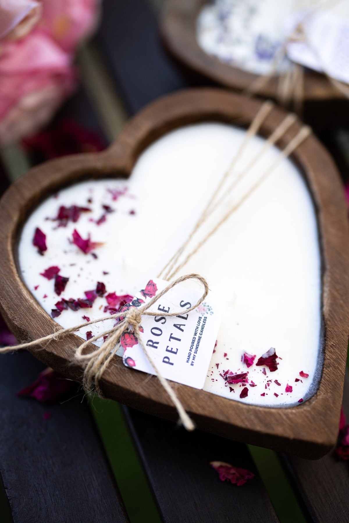 A heart-shaped wooden candle holder filled with white wax and decorated with dried rose petals, tied with twine and a small tag that reads 'ROSE PETALS'.