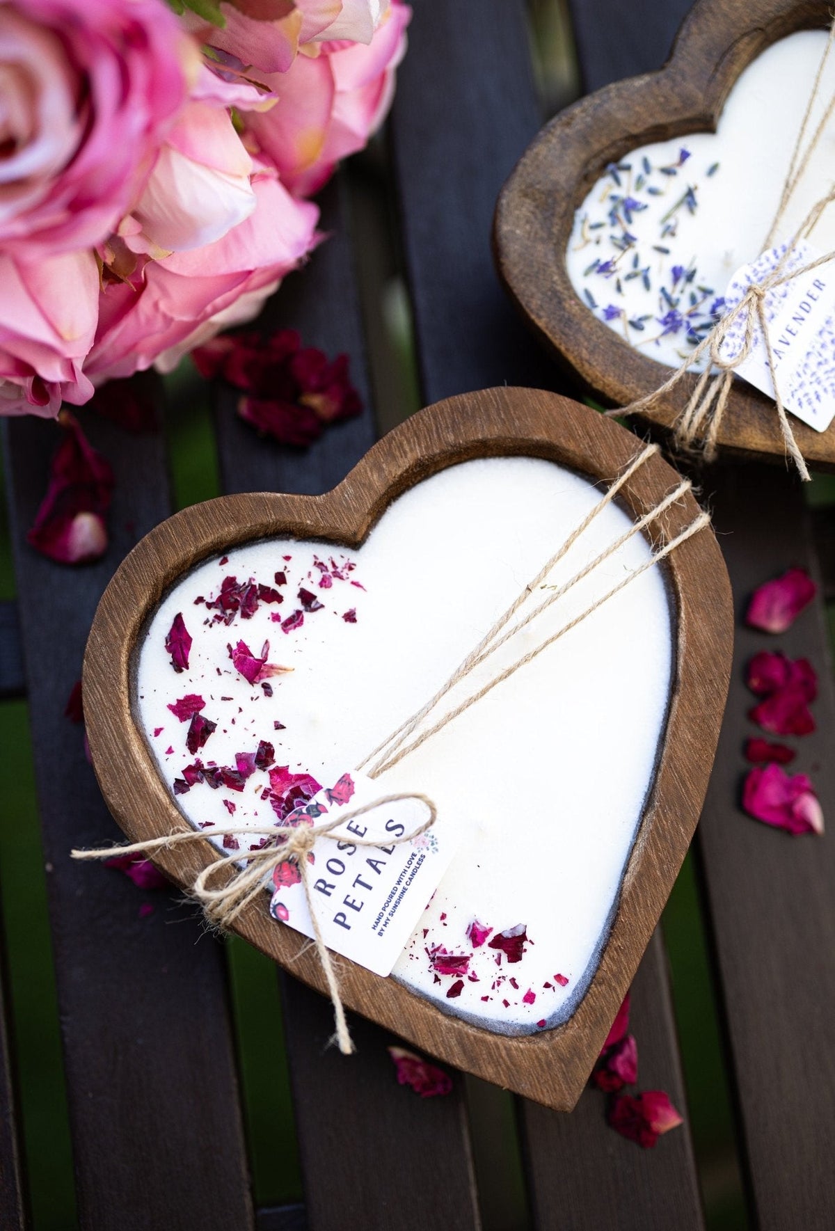 Two heart-shaped wooden soap dishes filled with white soap and dried flower petals, tied with twine and small labels reading ROSE PETALS and LAVENDER, displayed on a dark wooden slatted surface with pink roses nearby.
