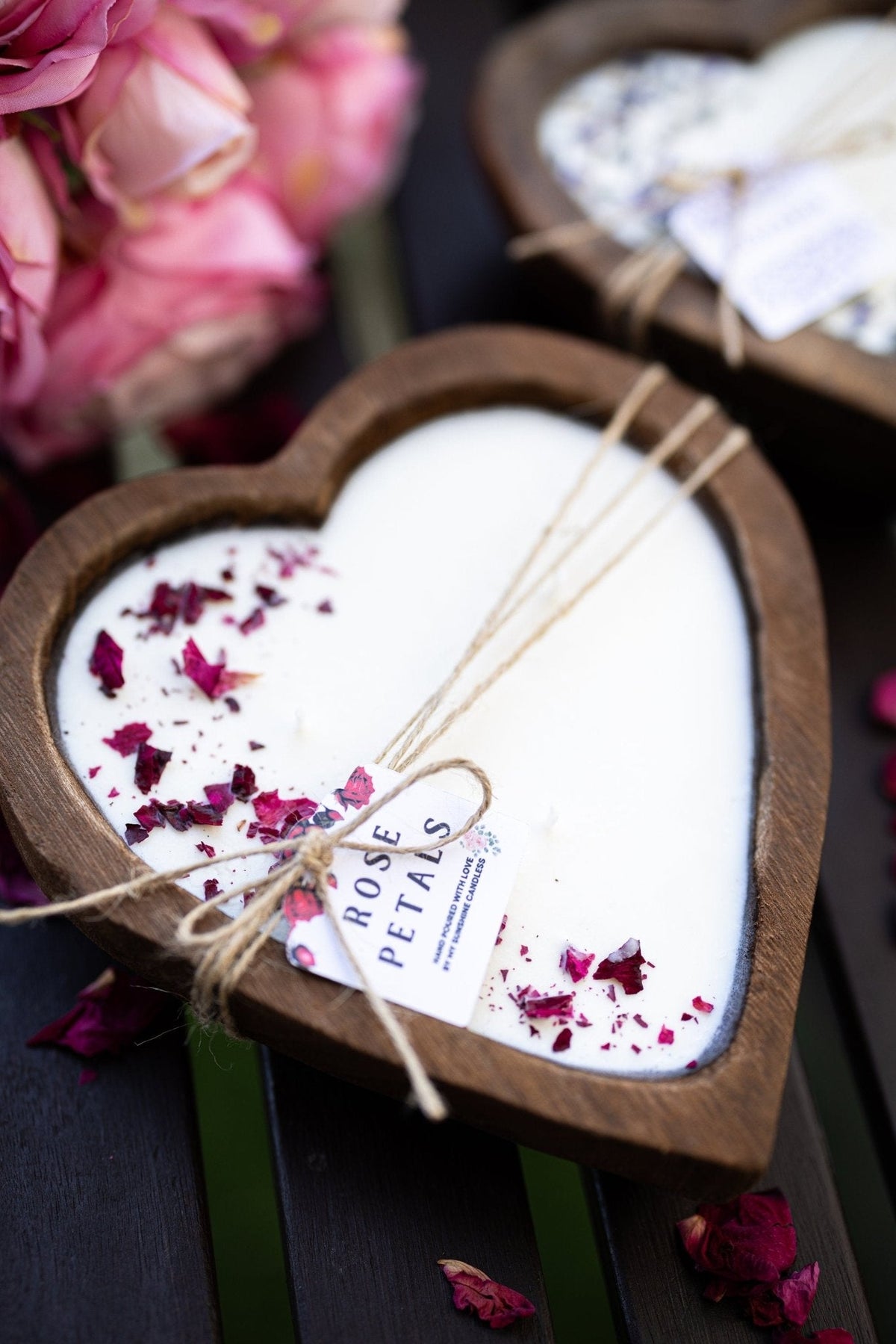 A heart-shaped wooden candle dish filled with white wax and decorated with dried rose petals, tied with twine and a small tag reading ROSE PETALS. Pink flowers and another similar candle are softly blurred in the background on a dark wooden surface.