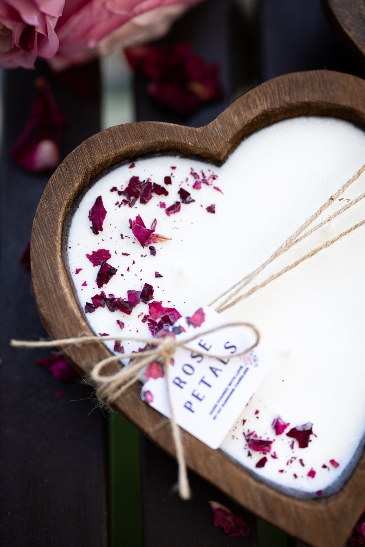 A heart-shaped wooden candle holder filled with white wax and decorated with dried rose petals, tied with twine and a small tag that reads 'ROSE PETALS'.