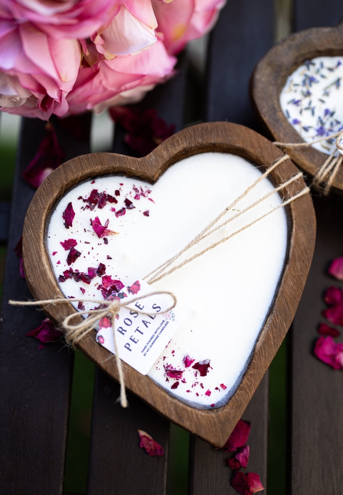 Heart-shaped wooden candle holder filled with white wax and decorated with dried rose petals, tied with twine and labeled 'ROSE PETALS', set on a dark wooden surface with pink flowers in the background.