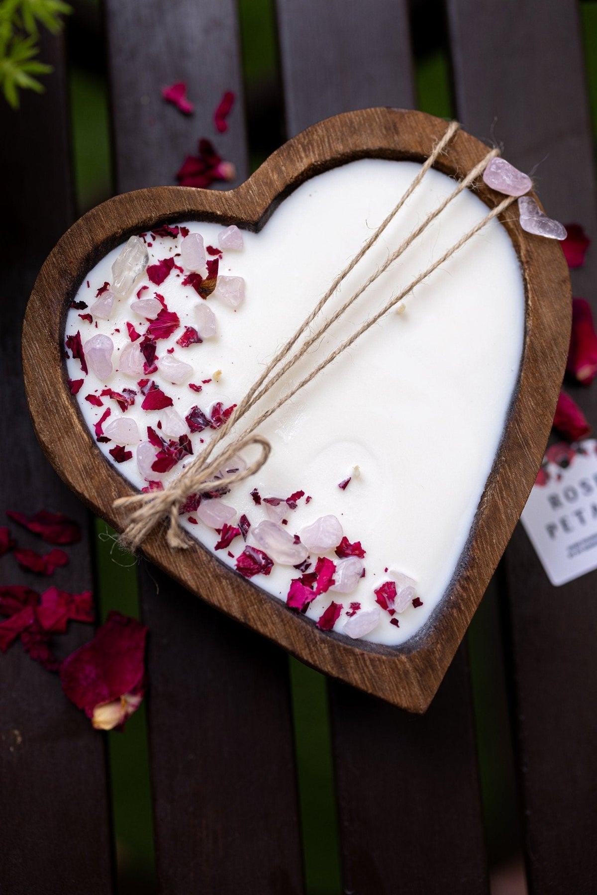 A heart-shaped wooden candle holder filled with white wax, decorated with pink rose petals and tied with twine, placed on a dark wooden surface.